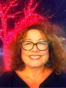 A smiling woman with blue glasses and shoulder-length brown curly hair wearing a black top against a pink light display blue glasses