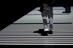 A horizontal crosswalk black and white striped being crossed by a pair of legs in vertical white and black stripes